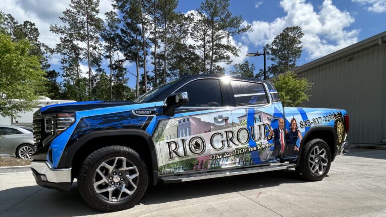 A blue pickup truck features a cityscape wrap and photos of two agents for the Rio Group real estate team.