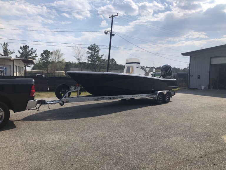 A matte black boat on a trailer hitched to a pickup truck in a parking lot.