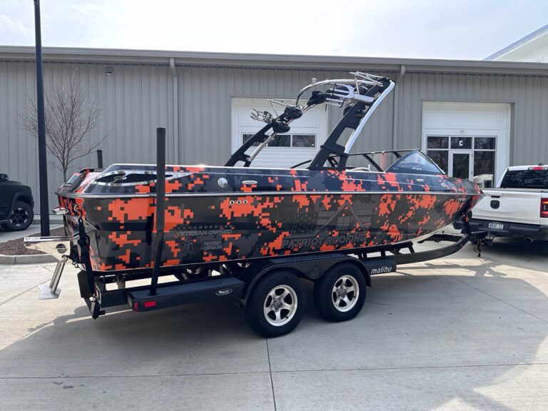 An orange and gray camouflage-wrapped boat advertising a roofing company sits on its trailer outside a warehouse.
