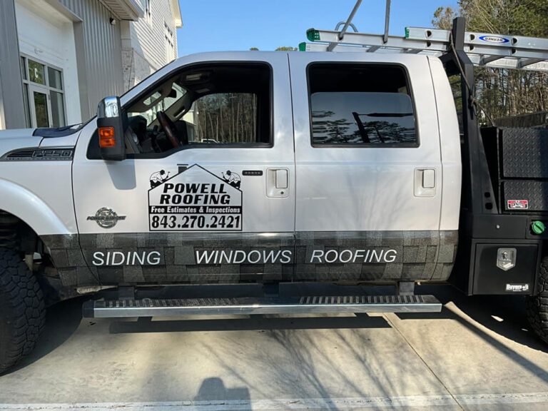 A white flatbed truck with Powell Roofing siding, windows, and roofing decals parked in a driveway.