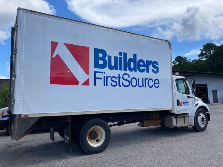 A white box truck displays large red and blue Builders FirstSource logo graphics on its side.