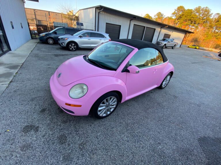 A matte pink vehicle wrap covers a Volkswagen Beetle convertible parked outside a commercial garage.