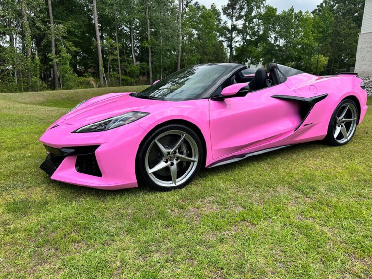 A glossy pink vehicle wrap covers a Corvette convertible parked on grass near a building.