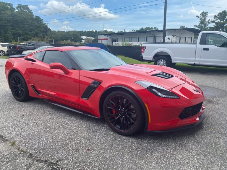 A glossy red Corvette with a gloss finish wrap sits parked in an outdoor lot near other vehicles.