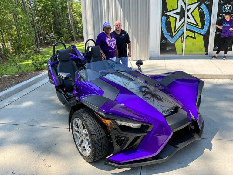 Two people stand beside a purple and black wrapped Polaris Slingshot outside the shop.