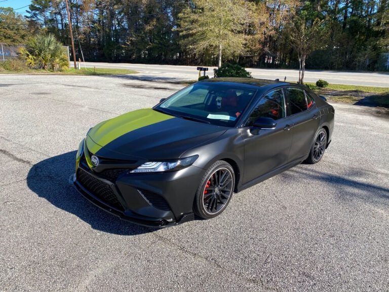 A matte black sedan with an olive green racing stripe wrap is parked on a street near a wooded roadside.