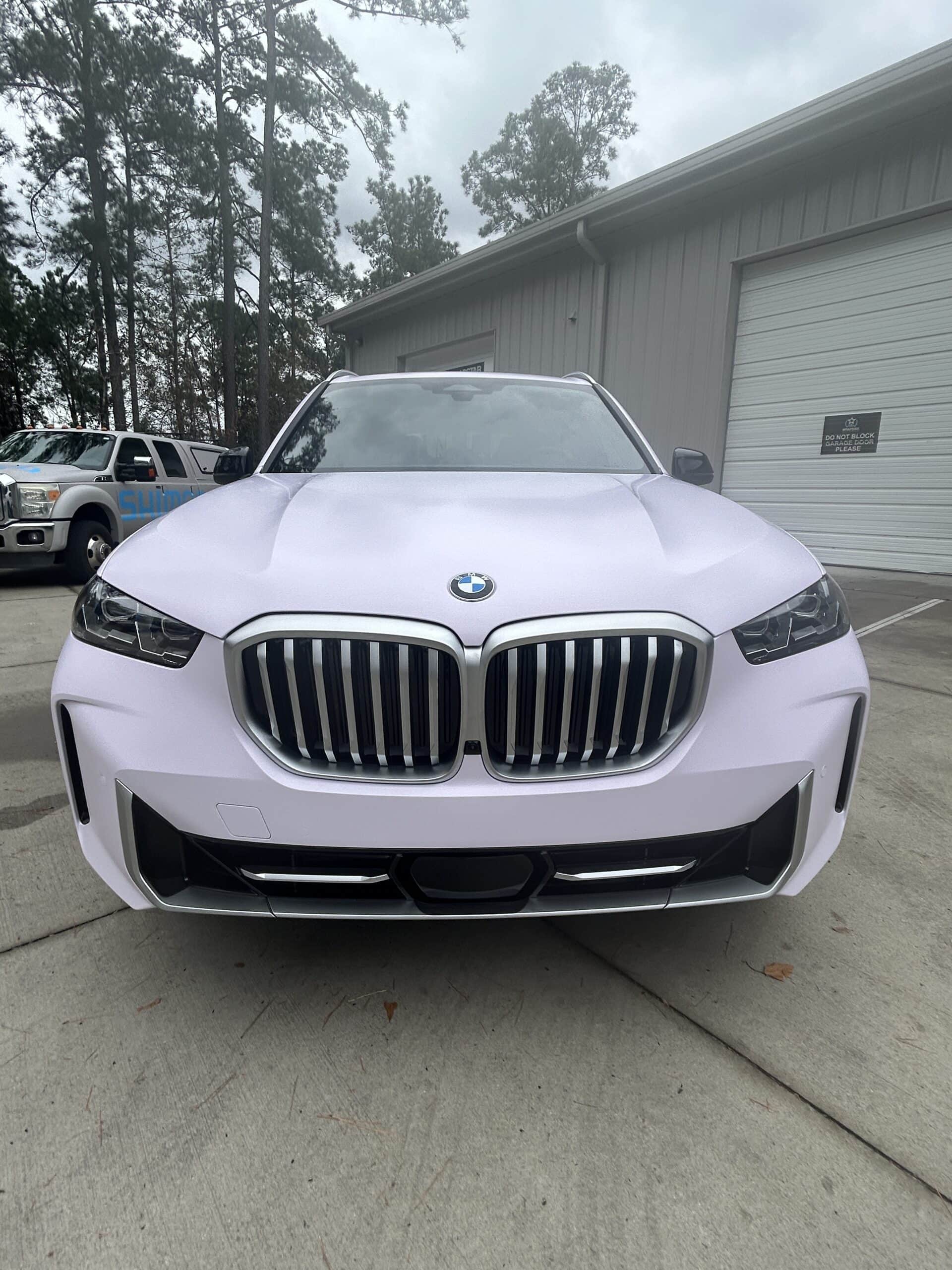 A white BMW SUV with a smooth color-change wrap, parked in front of a metal shop building.