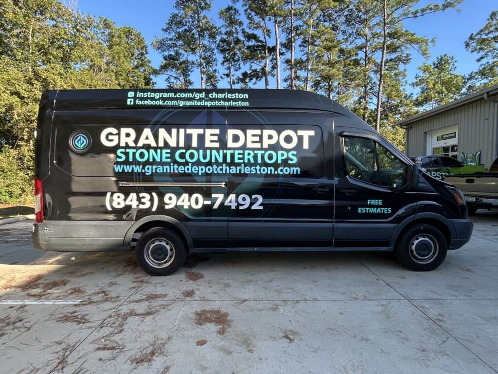 A black cargo van wrapped for Granite Depot Stone Countertops is parked near trees and a building.