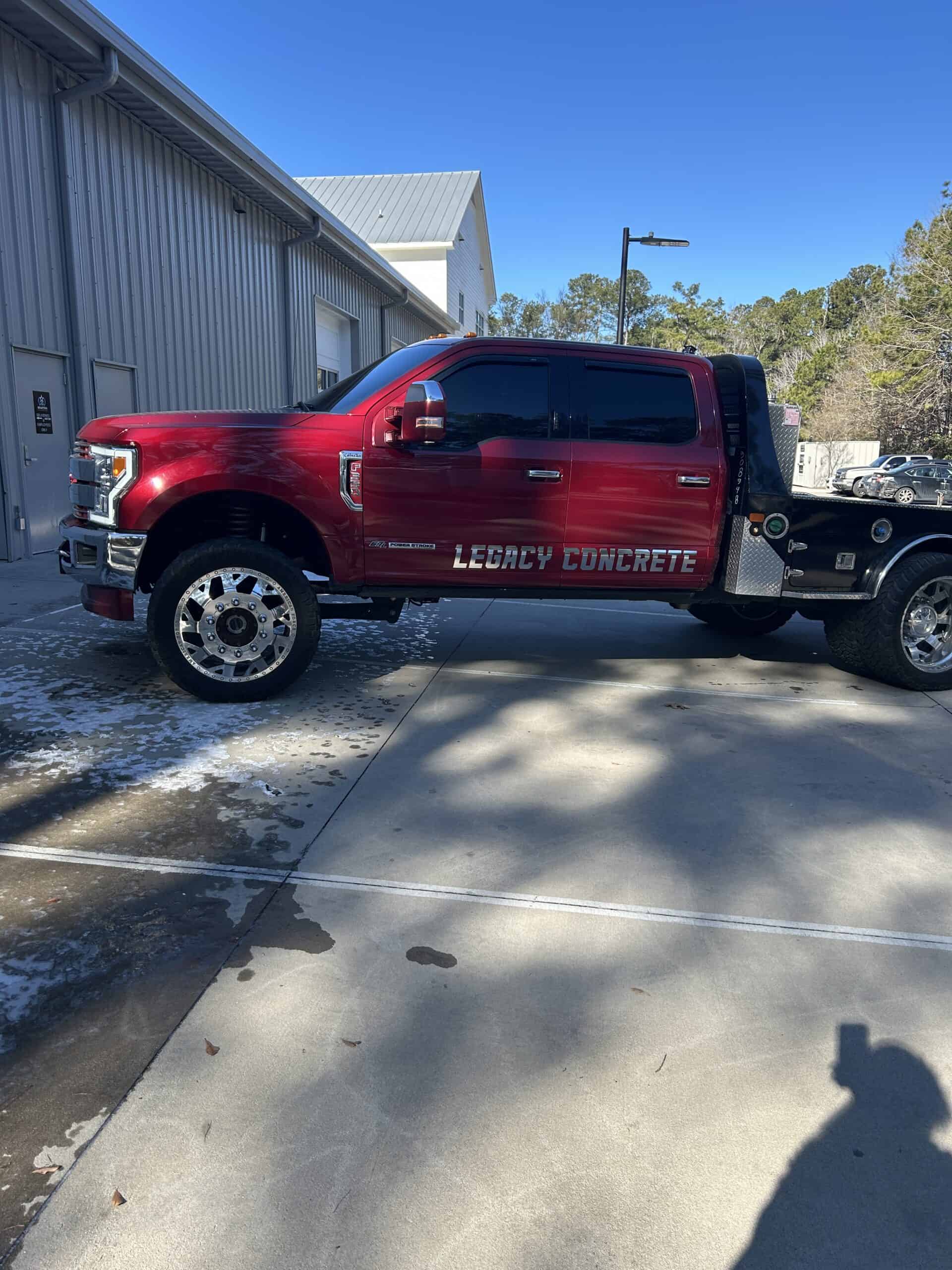 A red flatbed tow truck with chrome wheels displays a Legacy Concrete lettering wrap outside a shop.