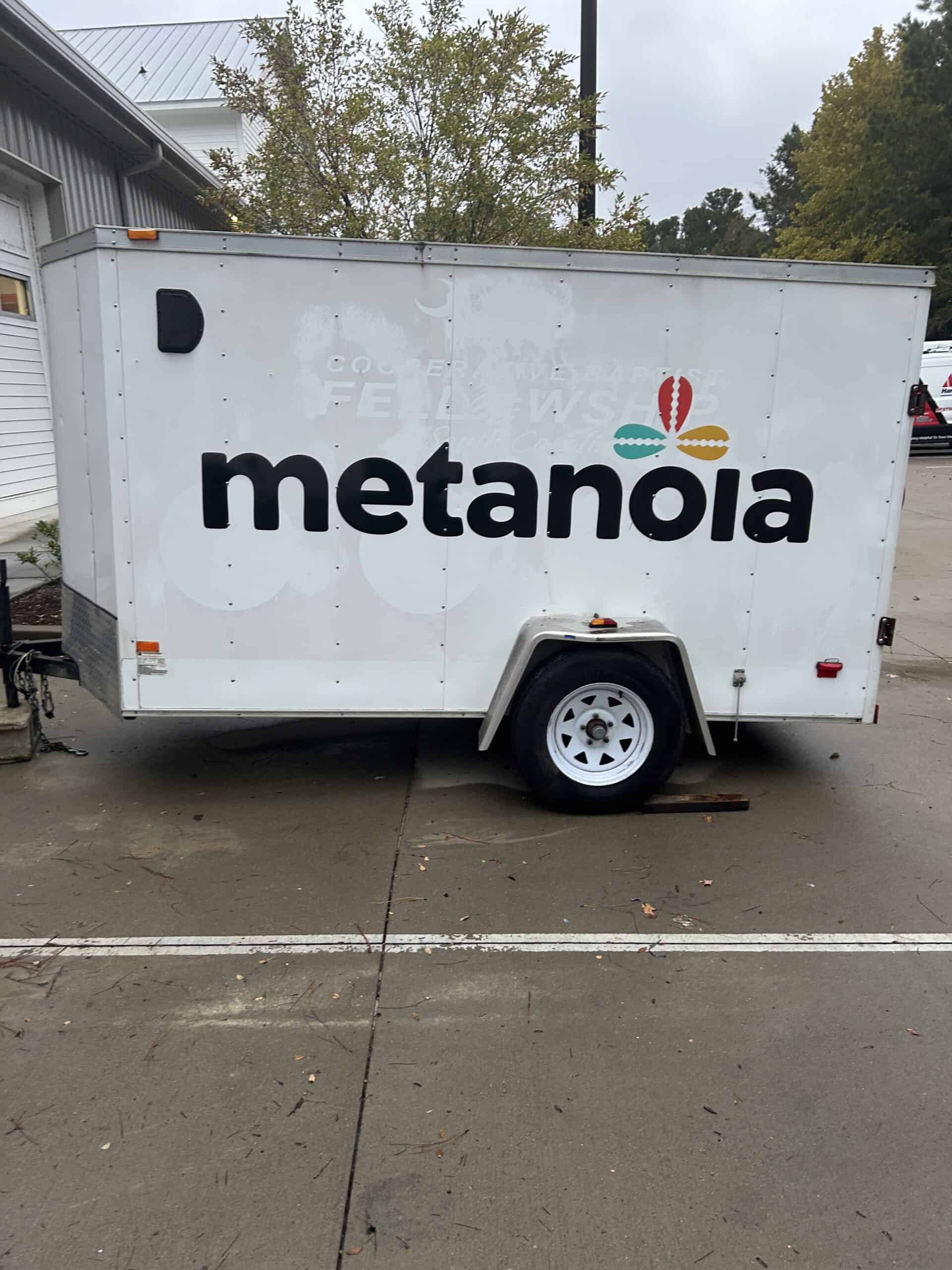 A white enclosed trailer wrapped with the Metanoia logo, with faint traces of an older design showing through.