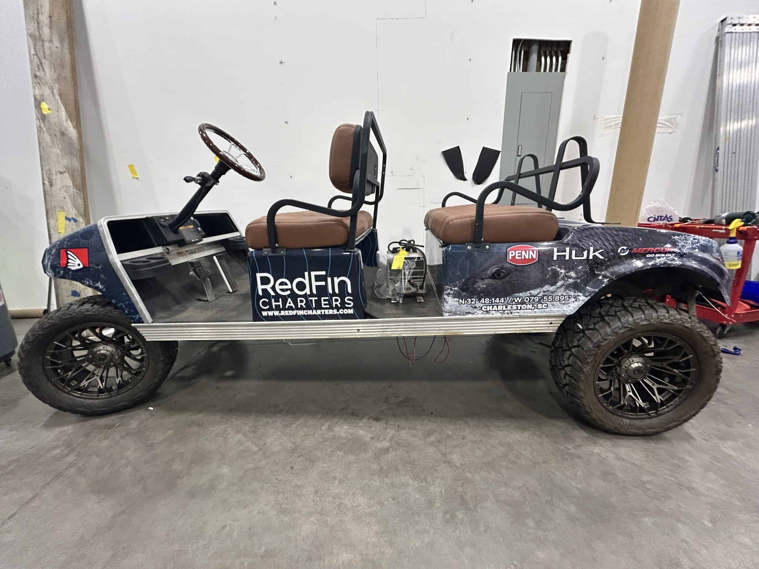 A golf cart chassis without body panels wrapped with RedFin Charters branding and sponsor logos in a workshop.