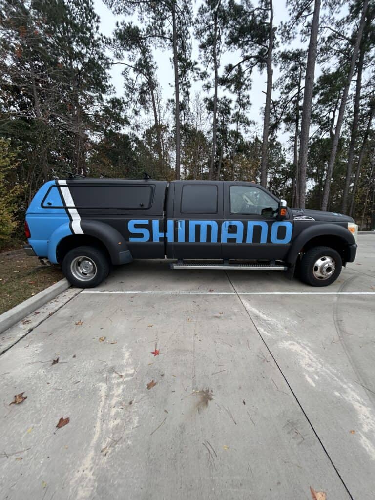A black dually pickup truck with a teal accent stripe displays a large Shimano logo wrap.