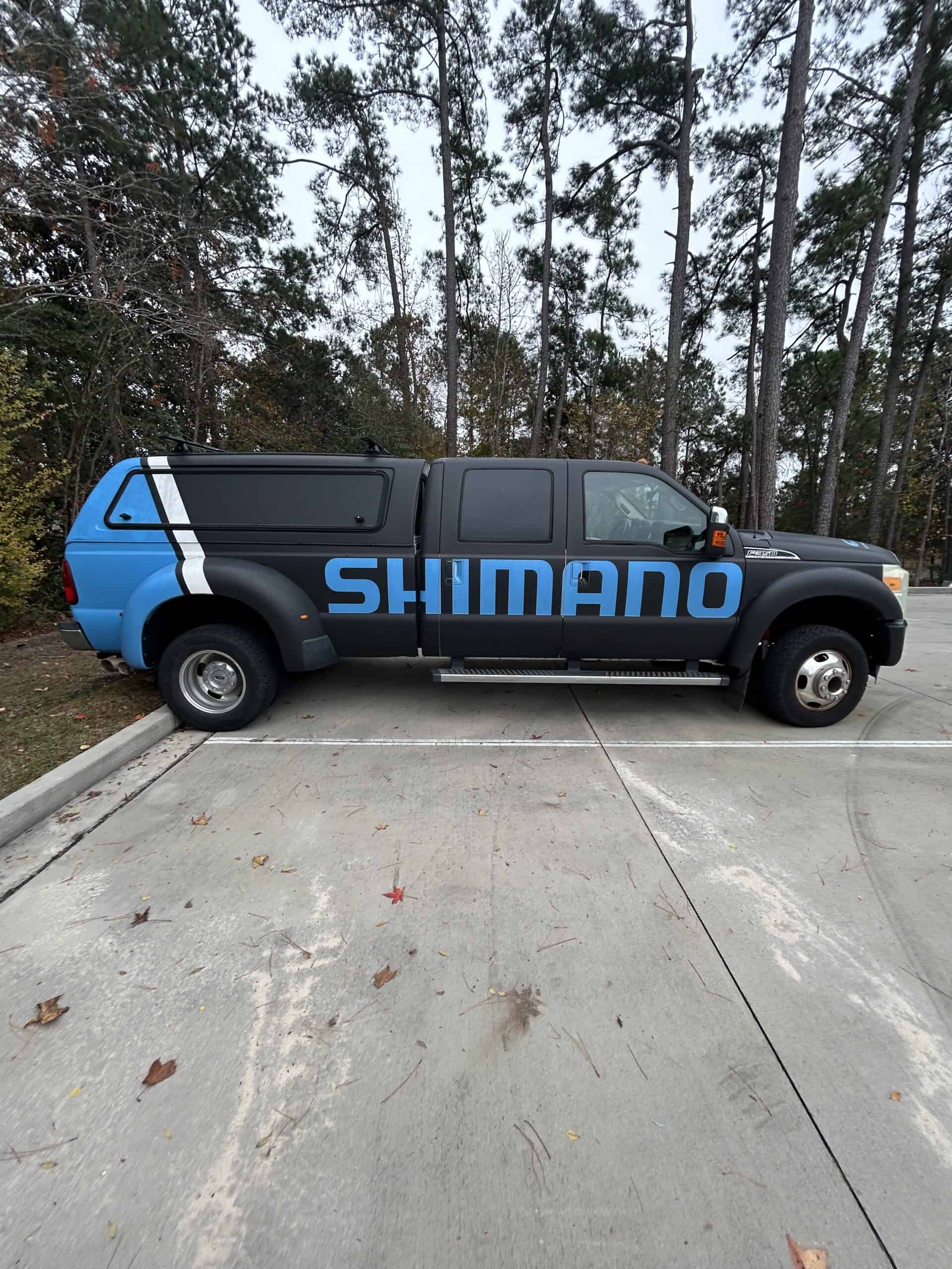A black dually pickup truck with a teal accent stripe displays a large Shimano logo wrap.