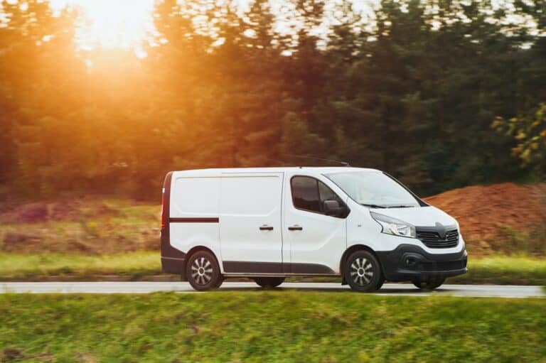 A white cargo van drives down a rural road as the sun sets behind trees.