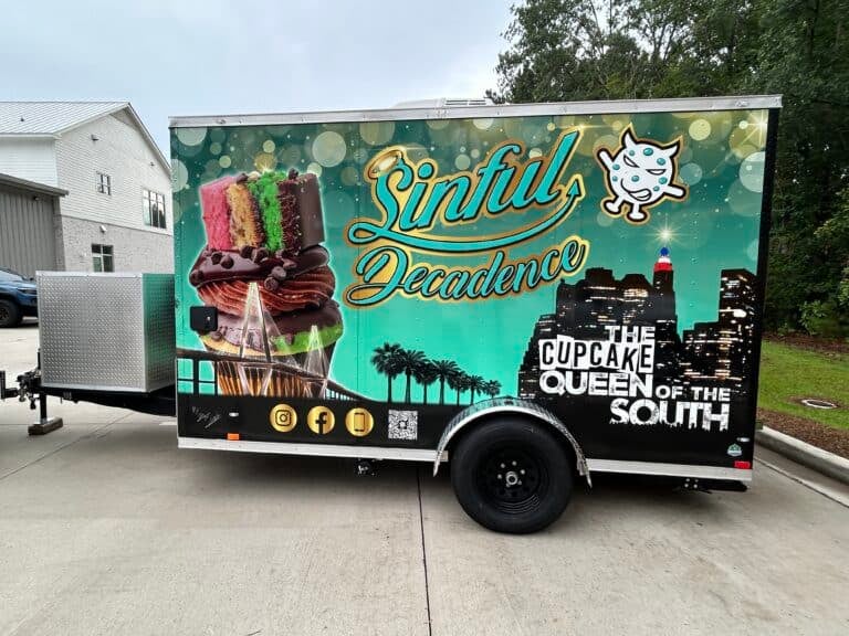 A food trailer wrapped in teal and gold for Sinful Decadence, The Cupcake Queen of the South, parked outside a shop.